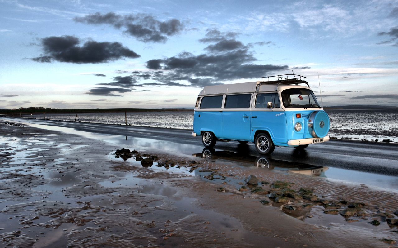 Vintage blue campervan on a coastal road beside the beach under a moody sky.