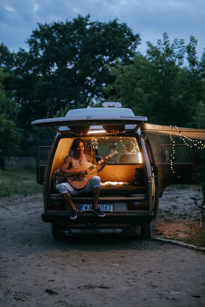 A young man sits in a van playing guitar surrounded by nature at dusk.